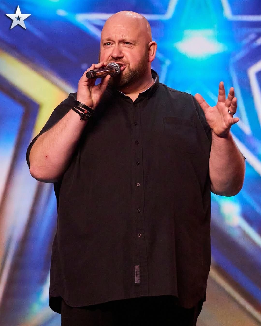 Bald man singing into a microphone on the Britain's Got Talent stage under blue star lighting.