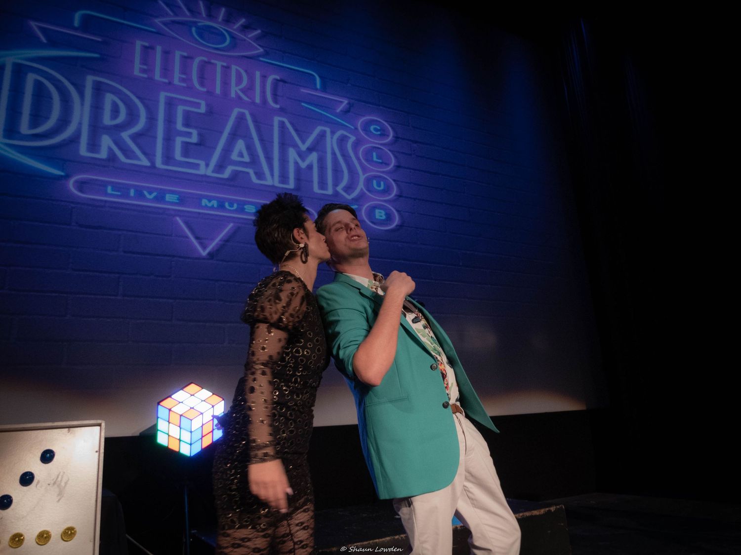 Two performers singing together on stage with blue neon 'Electric Dreams' sign visible.