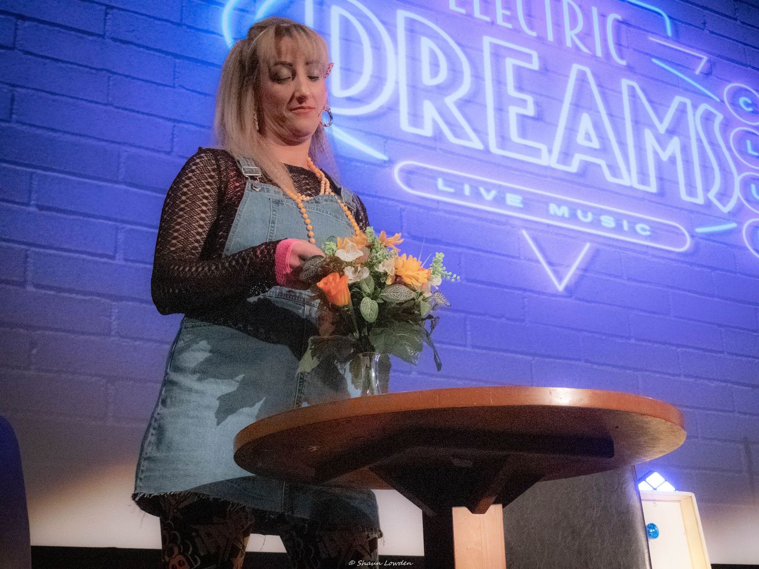 Performer in denim dress arranging flowers on stage with neon signage.