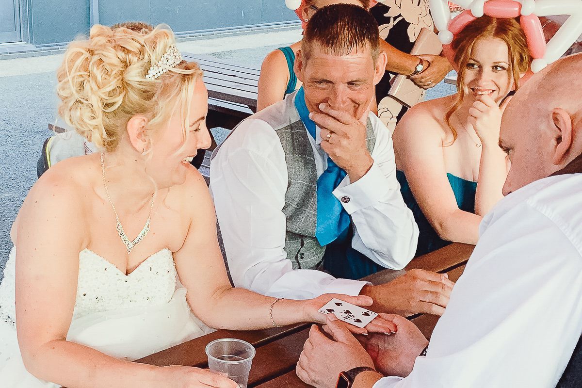 A magician performs a card trick for a laughing bride and groom at a wedding reception.