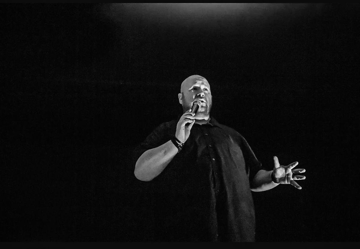 Bald man in black shirt singing into microphone on dark stage in black and white performance photo.