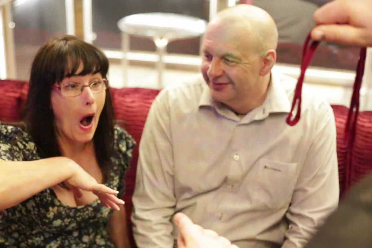 A surprised woman and bald man sit on red chairs as someone presents a red item to them.