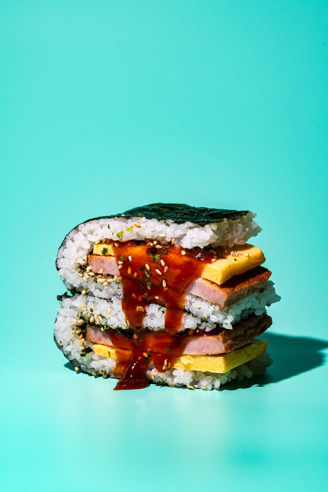 Stacked sushi rolls with rice, nori, ham, egg, and red sauce on a teal background.