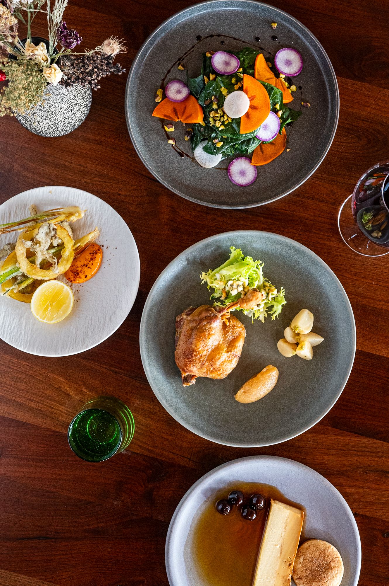 Overhead view of elegant restaurant dishes on wooden table featuring roasted chicken, colorful vegetable salad, and dessert plates.