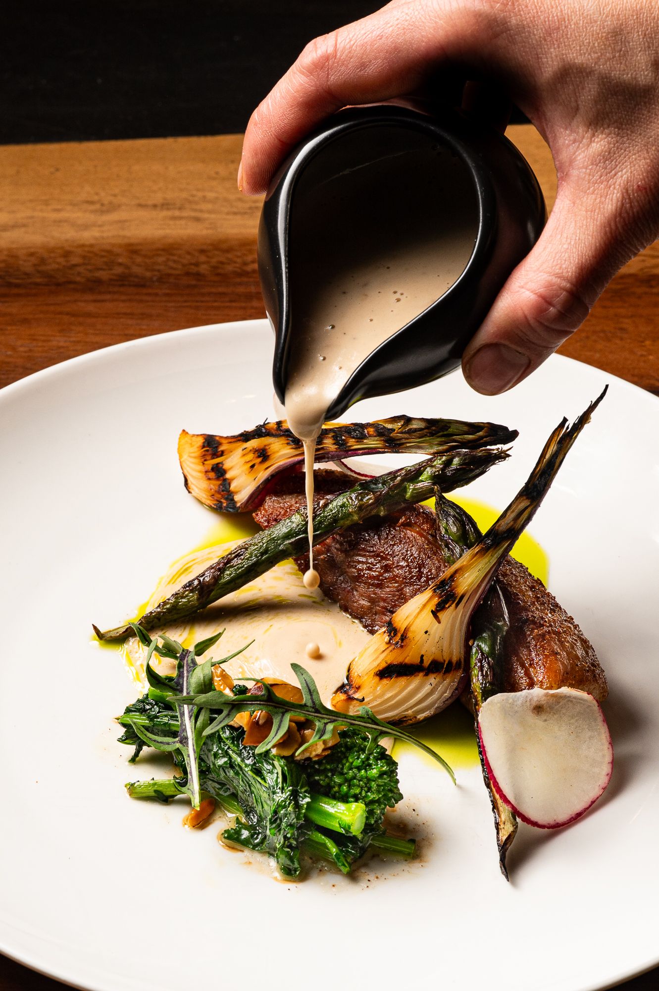 Chef pouring creamy sauce over grilled lamb chops with asparagus, radish, and greens on a white plate.