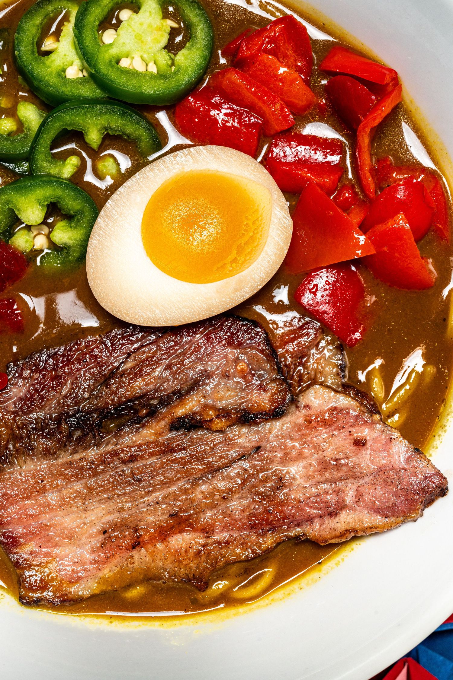 A bowl of ramen with grilled beef, soft-boiled egg, red peppers, and sliced jalapeños in a rich broth.