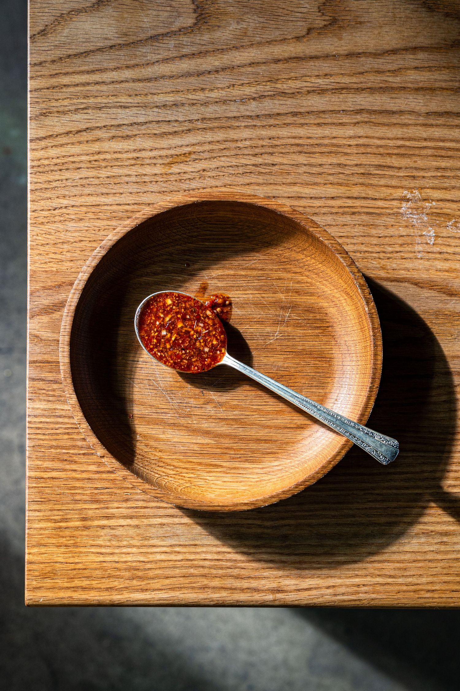 A spoonful of red chili flakes resting in a small wooden bowl on a light oak wooden surface.
