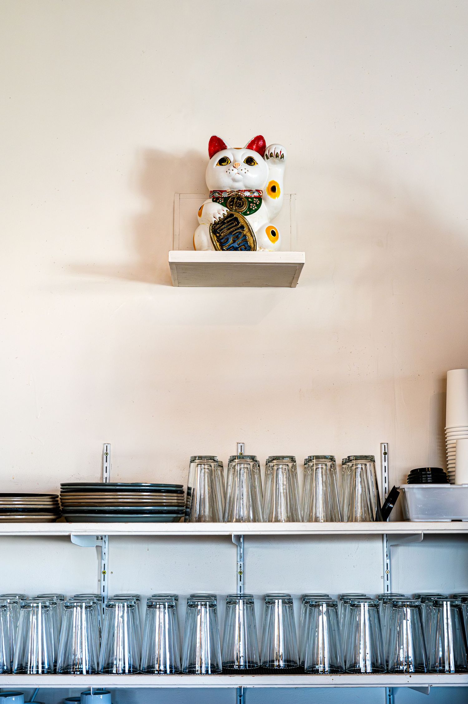 Maneki-neko lucky cat figurine on wall shelf above restaurant bar shelves stocked with clear drinking glasses and plates.