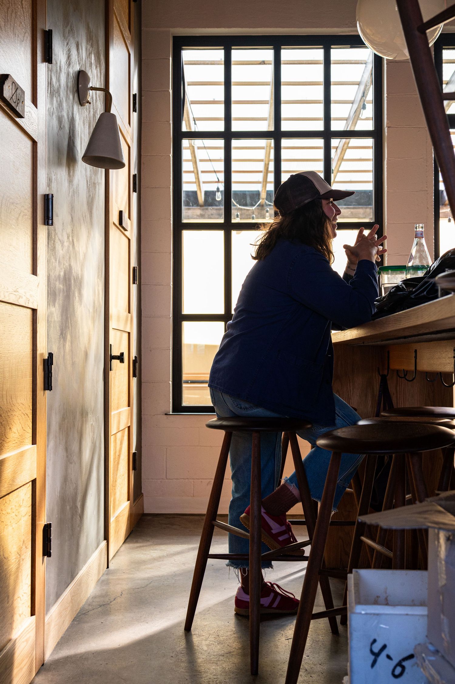 Woman sitting on a wooden stool at a counter by a large window in a cozy cafe, wearing a blue jacket and cap.