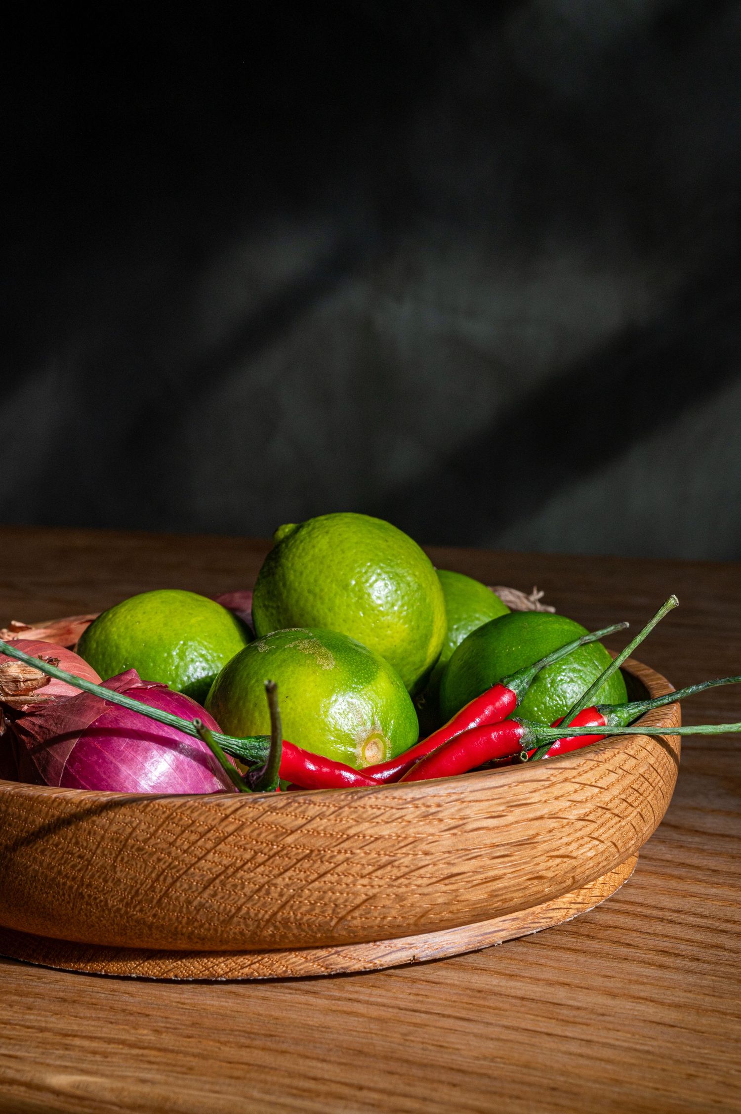 Wooden bowl filled with fresh limes, red chili peppers, and red onion on a rustic wooden table with dark background.
