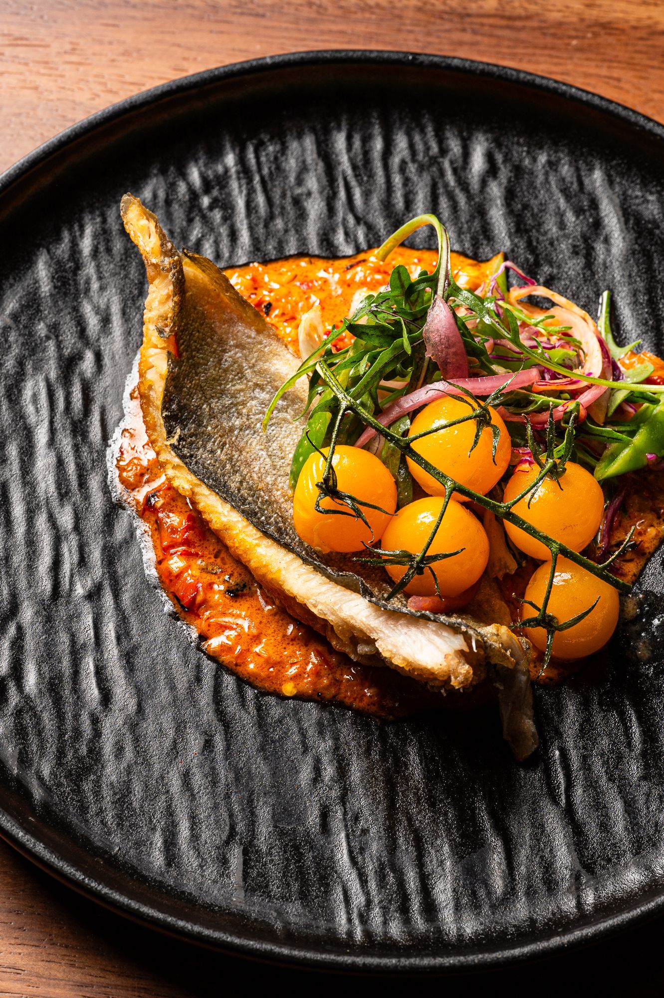Pan-seared fish fillet on tomato sauce with cherry tomatoes and fresh microgreens on a black textured plate.