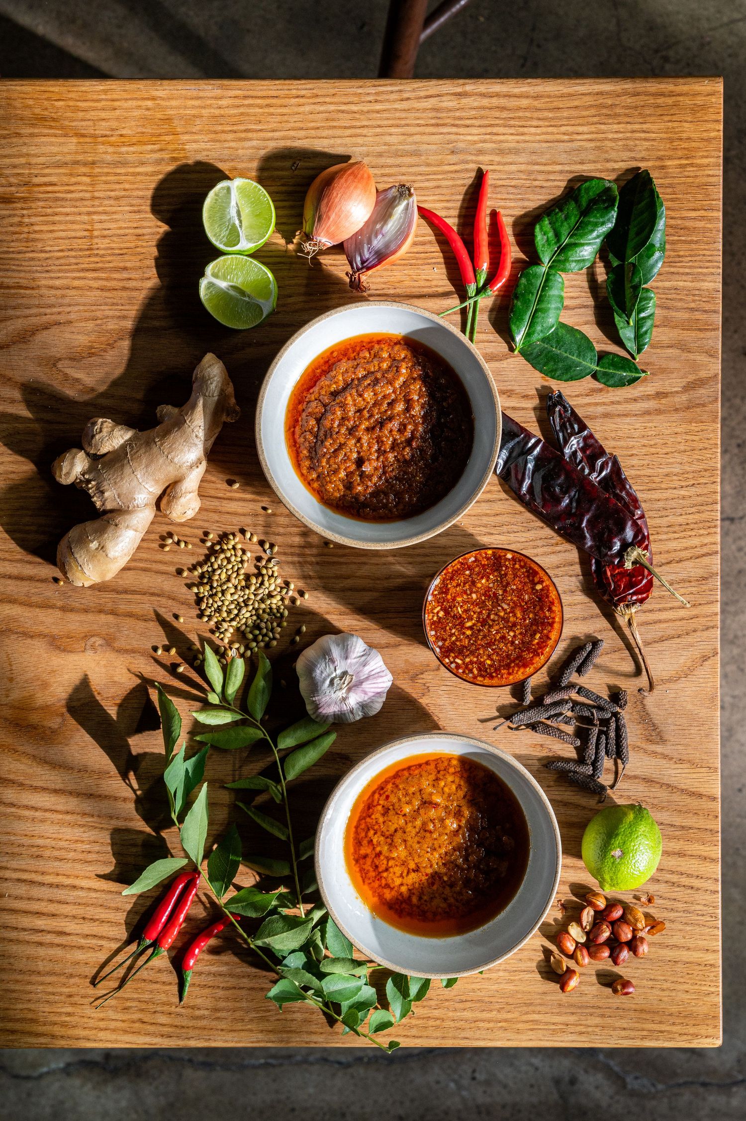 Two bowls of spicy chili paste surrounded by fresh ingredients including ginger, chilies, lime, garlic, and herbs on a wooden board.