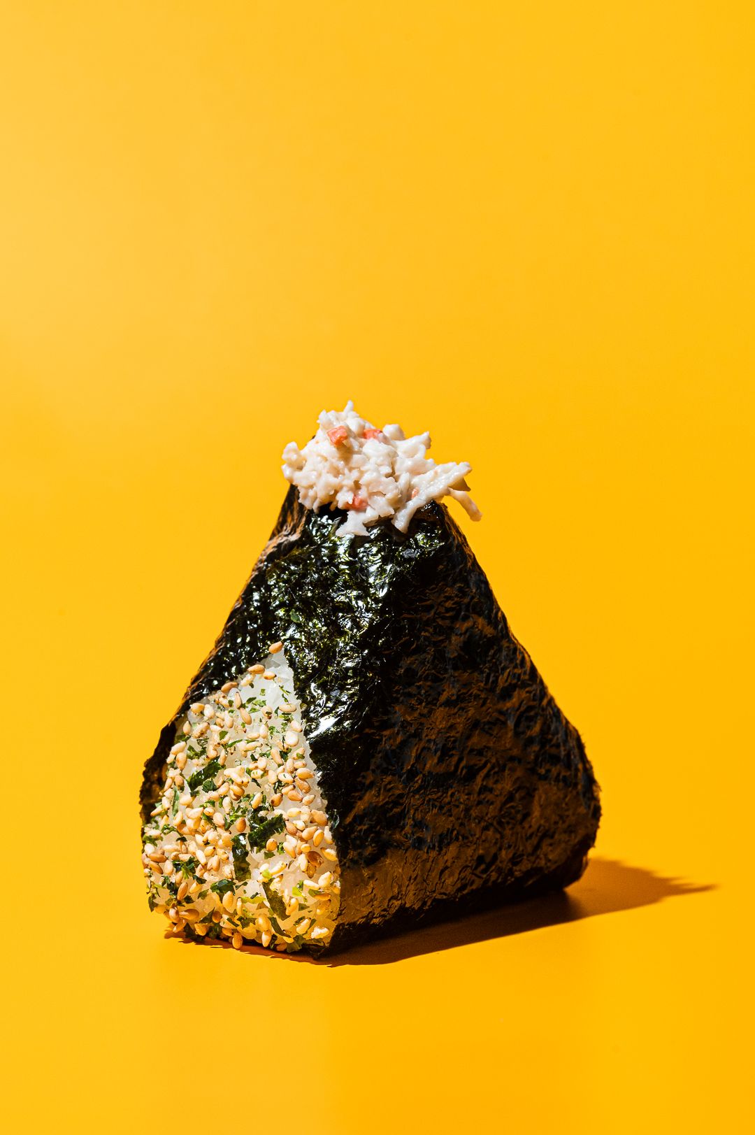 Japanese onigiri rice ball wrapped in nori seaweed with tuna topping on bright yellow background.