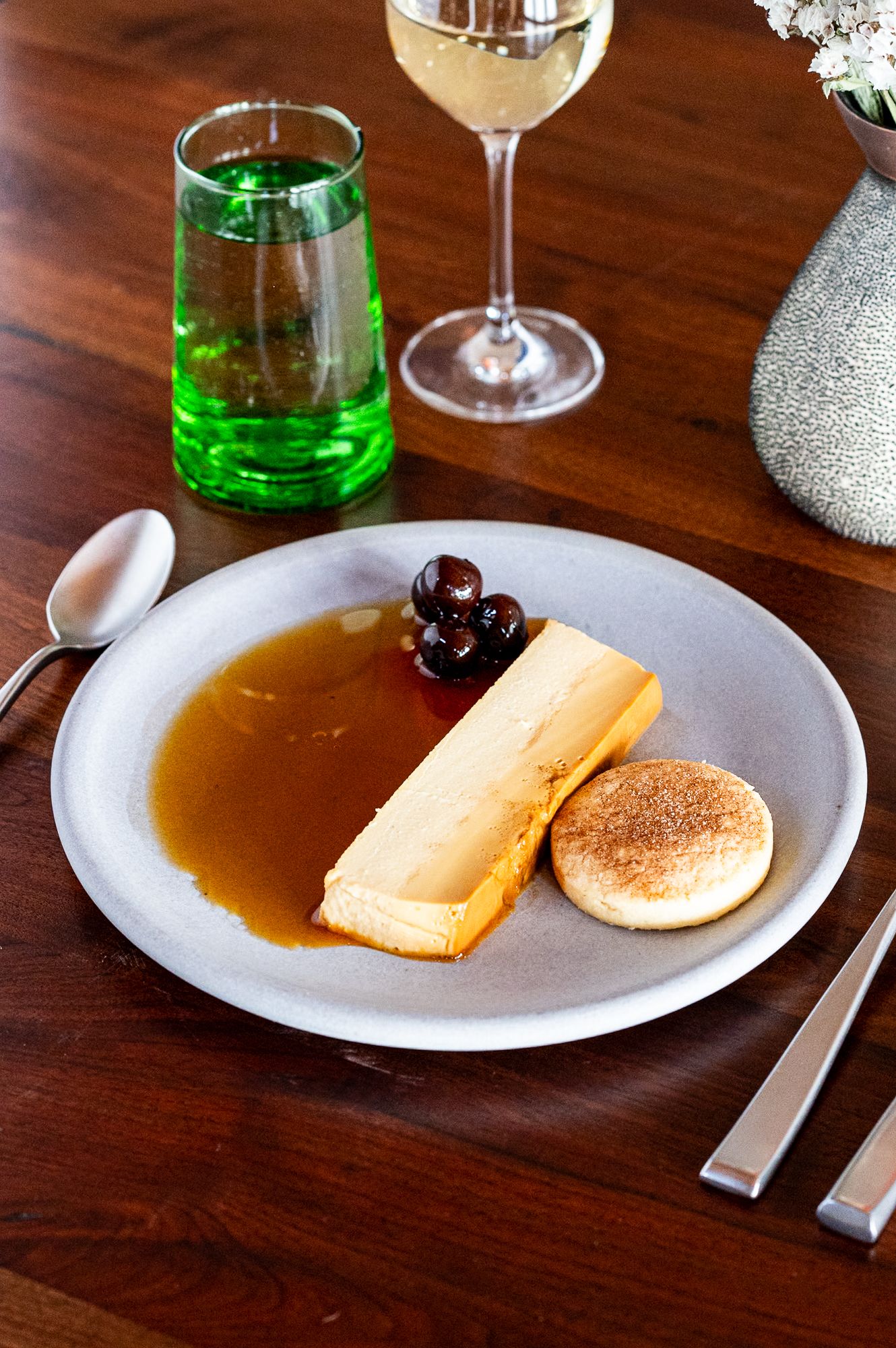 Elegant dessert plate with caramel flan, dark cherries, and a round cookie, served with white wine and green water glass.
