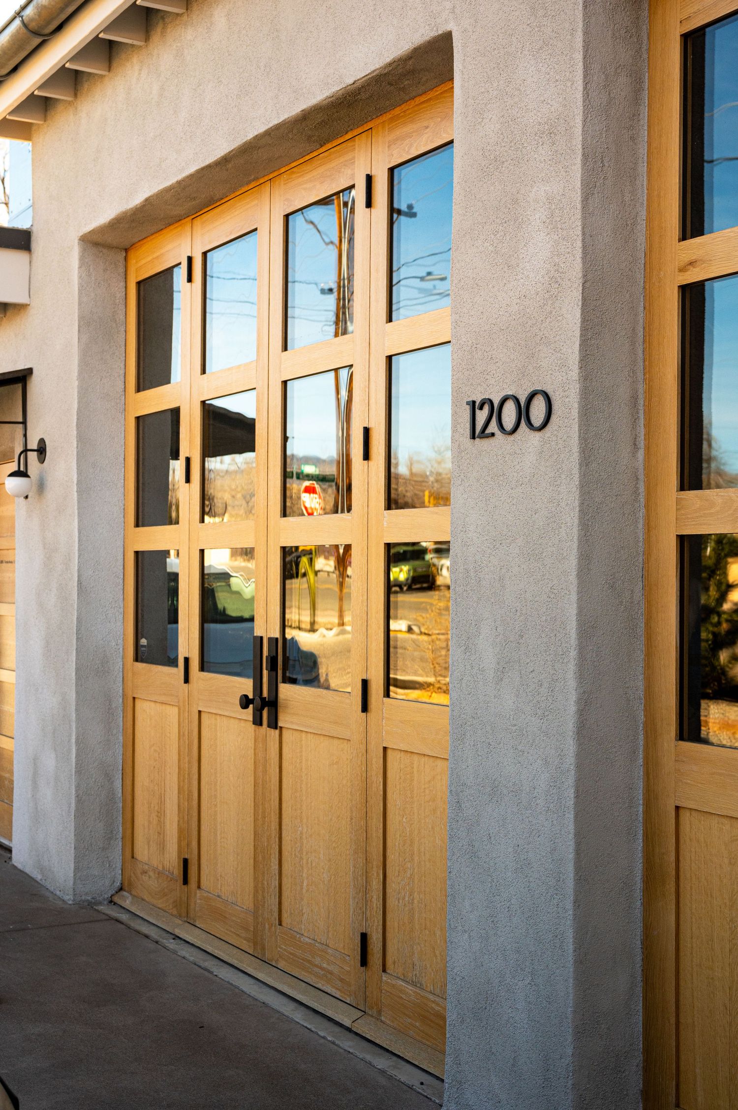 Modern wooden glass-panel entrance doors at address 1200, set in a stucco building facade, reflecting golden sunlight.