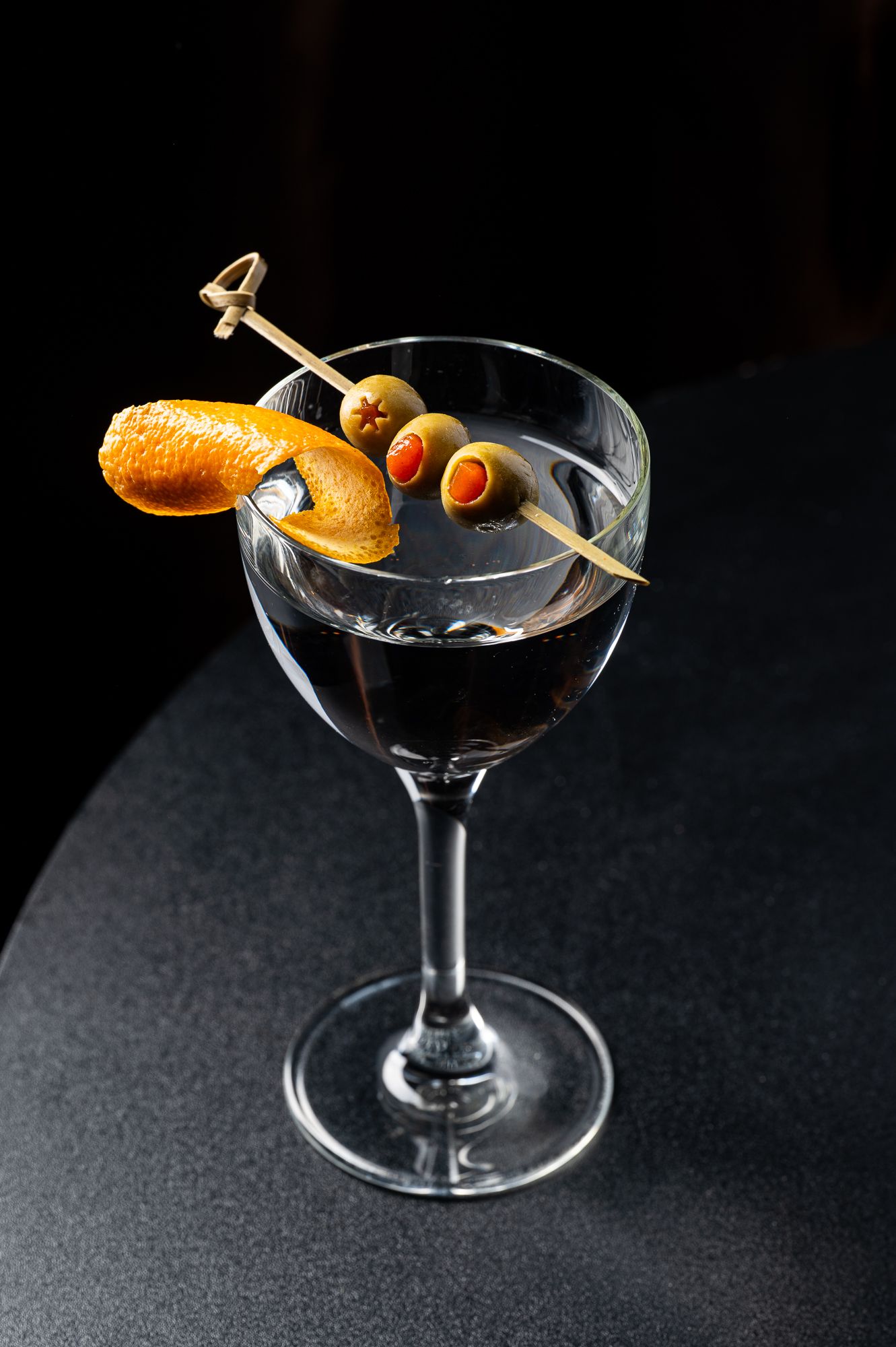 Elegant martini glass with clear cocktail, garnished with green olives on a skewer and a twisted orange peel, on dark surface.