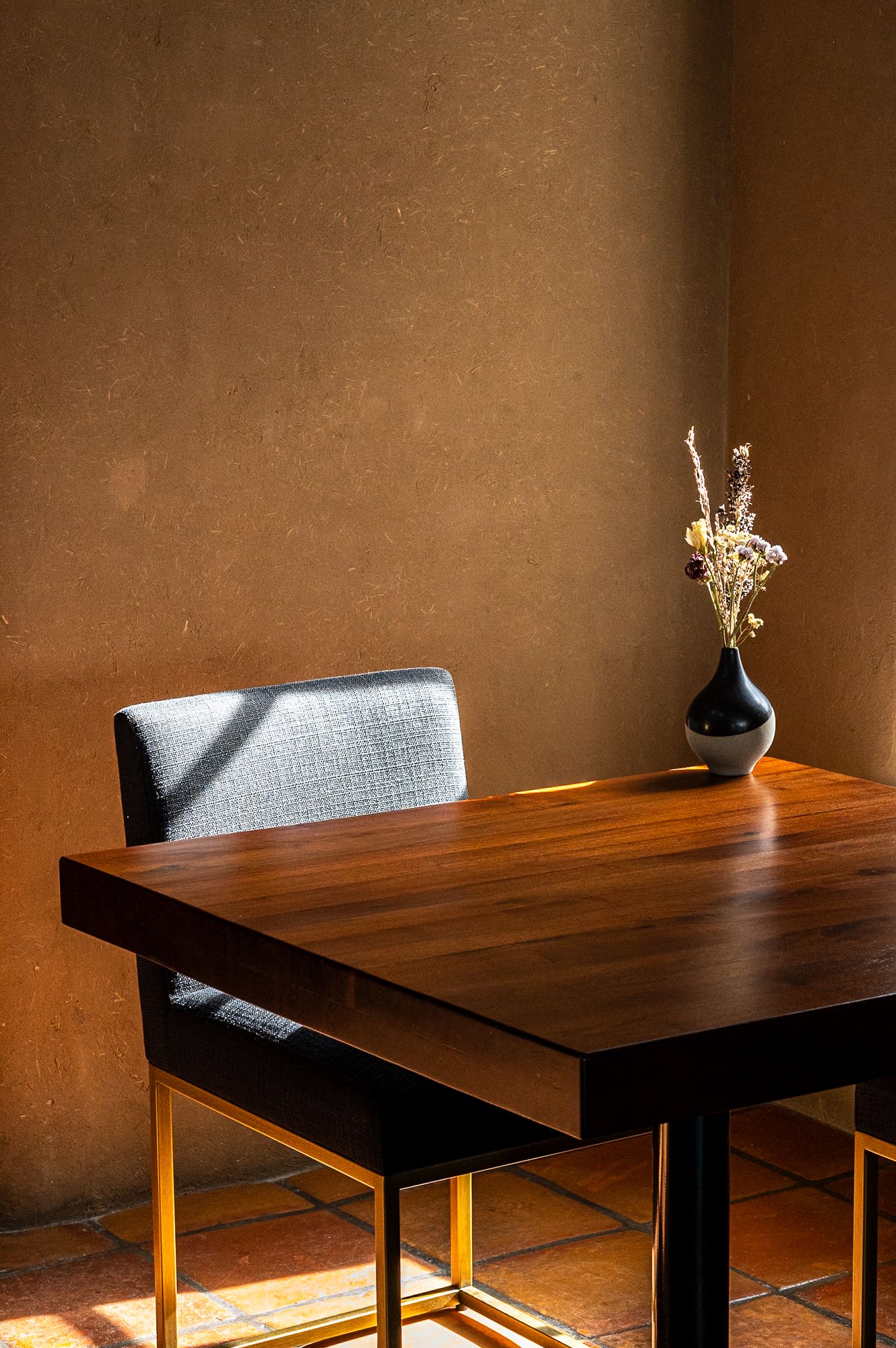 Sunlit wooden dining table with a black vase of dried flowers and a gray upholstered chair against a warm beige wall.