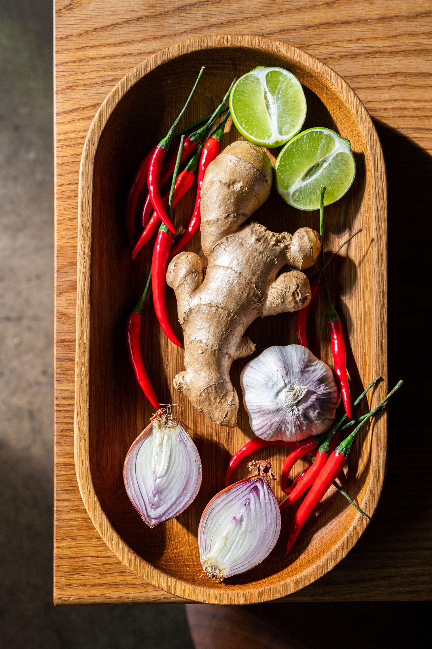 Wooden bowl filled with fresh ginger, garlic, red chili peppers, sliced lime, and halved red onion on a wooden surface.
