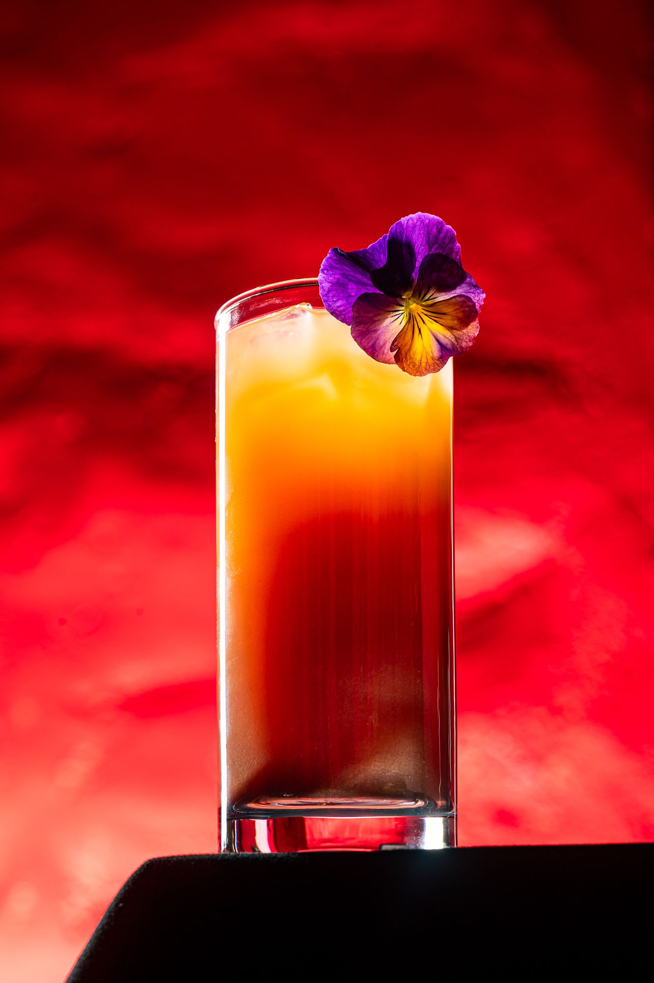 A vibrant orange cocktail garnished with a purple pansy flower in a tall glass against a red background.