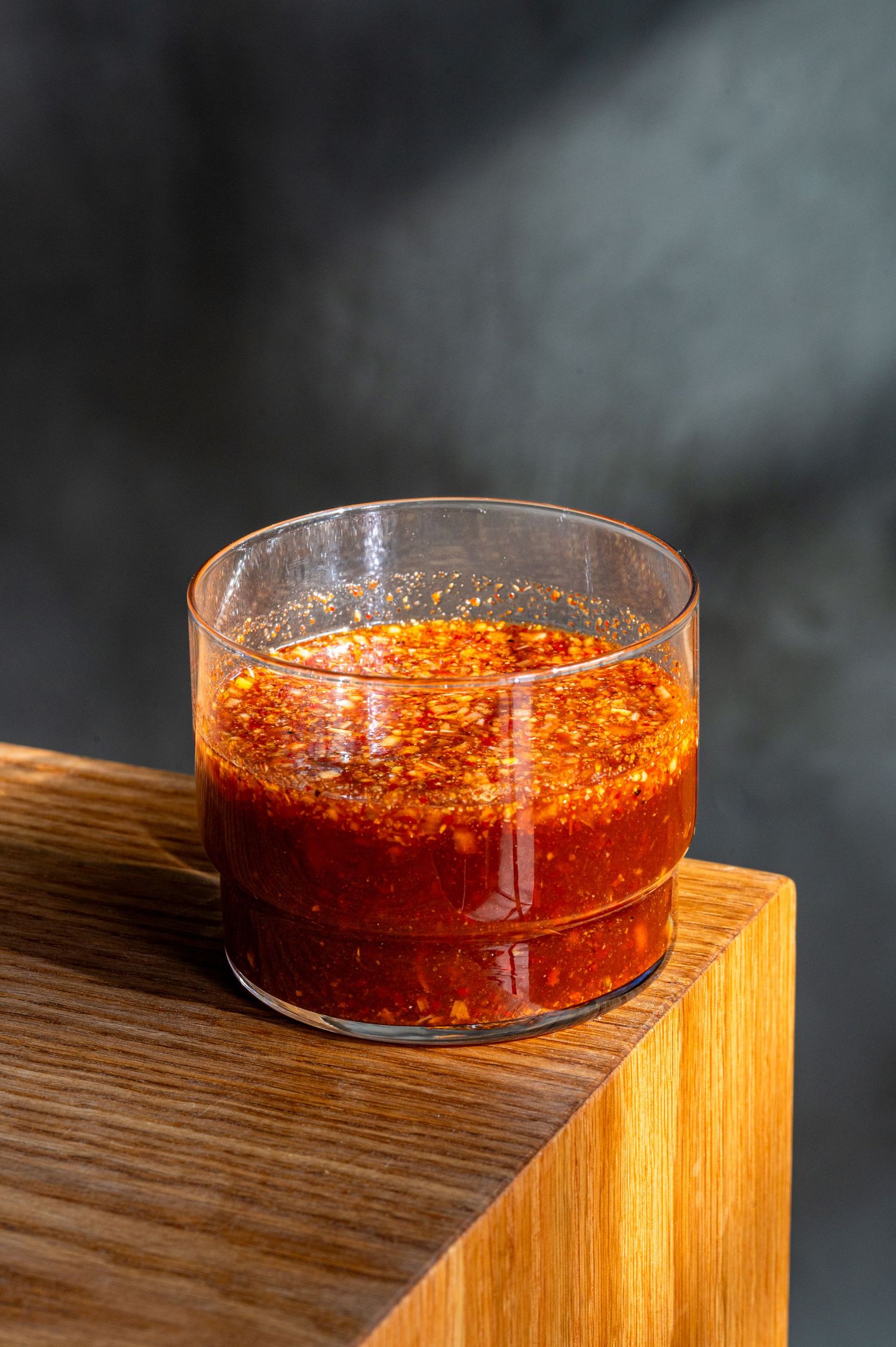A glass of vibrant red chili sauce with spices sits on a wooden surface against a dark background.