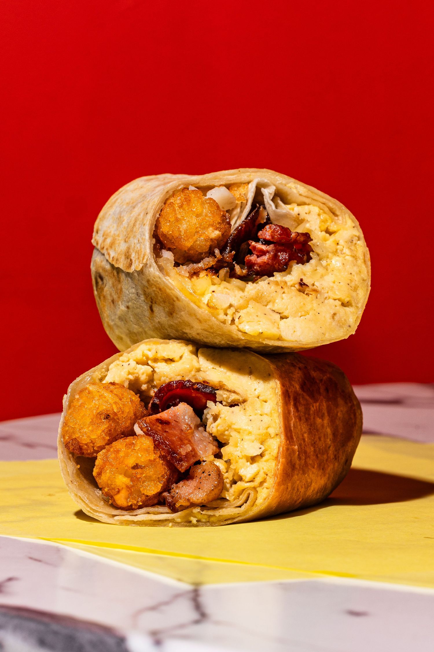 A halved breakfast burrito stacked showing scrambled eggs, bacon, tater tots, and fillings on a marble surface.