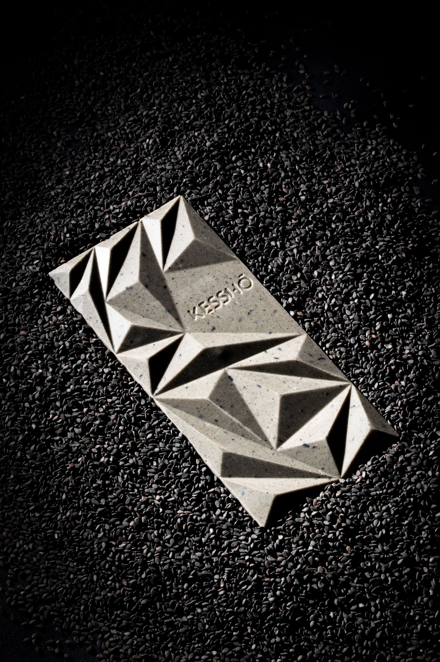 A geometric patterned KESSHO chocolate bar with angular facets resting on dark textured surface in dramatic black and white.