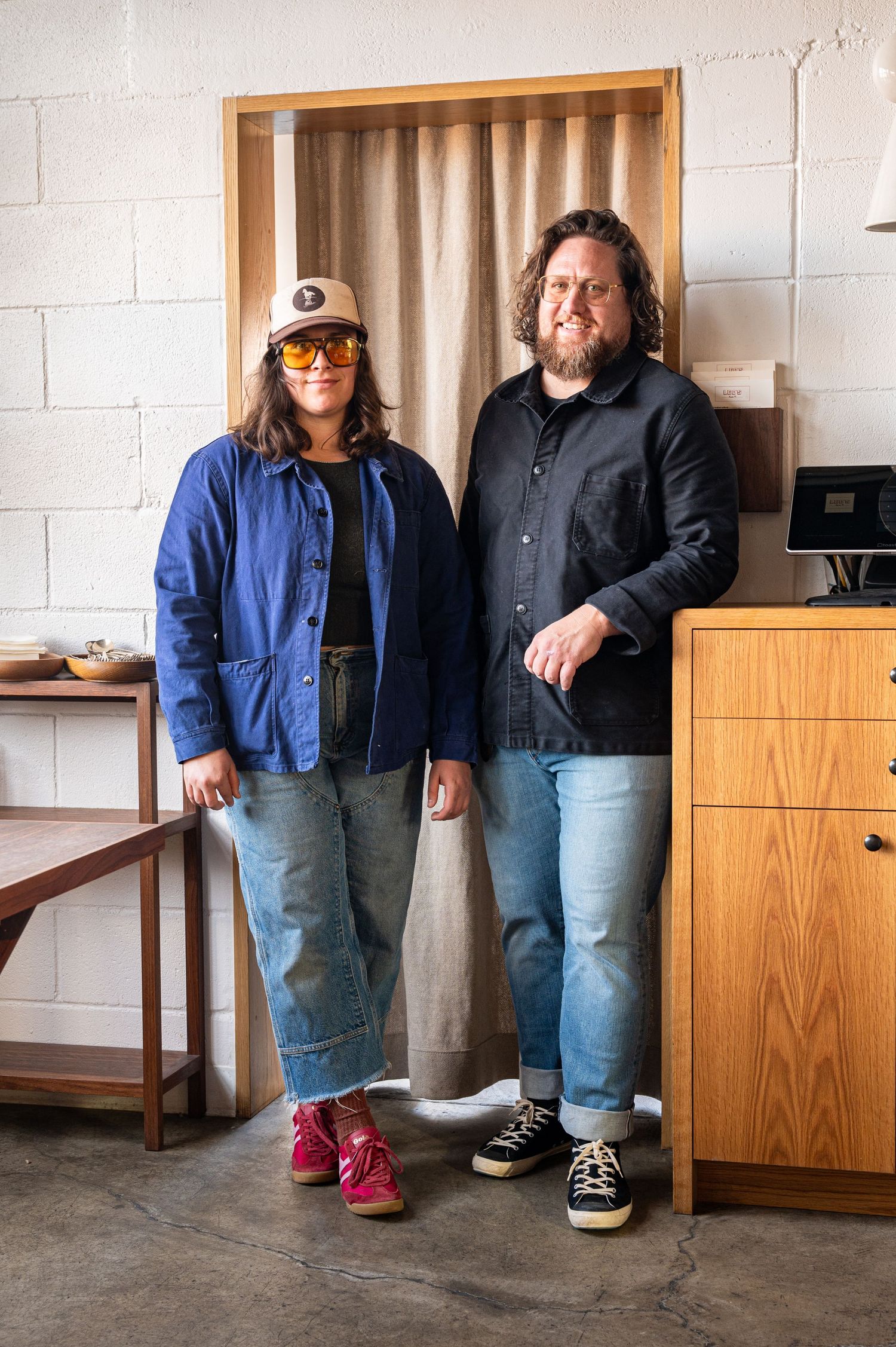Two people in casual denim outfits stand together in a modern workshop studio with wooden furniture.