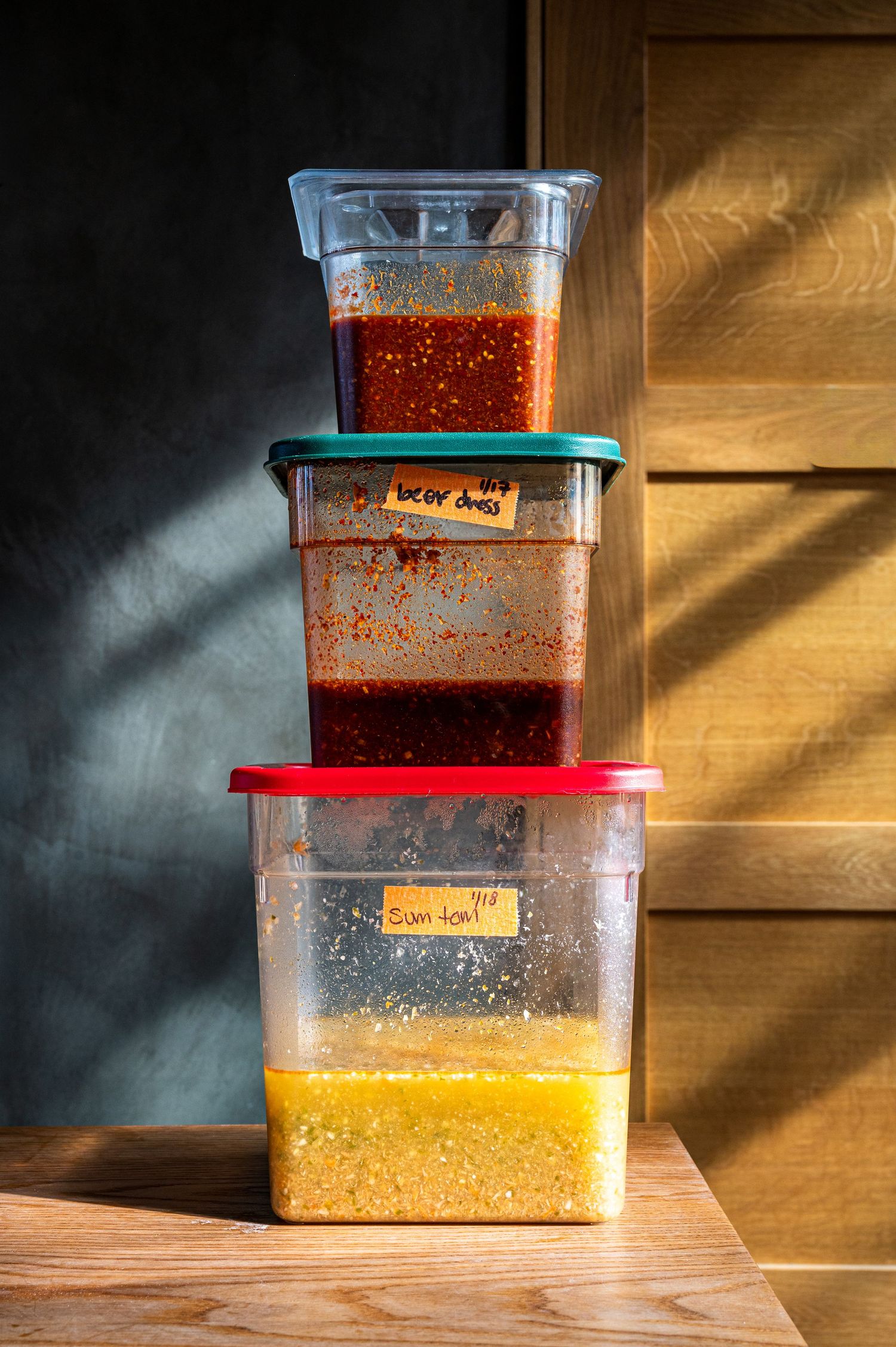 Three stacked clear food containers with red and green lids holding colorful sauces and grains on a wooden surface.