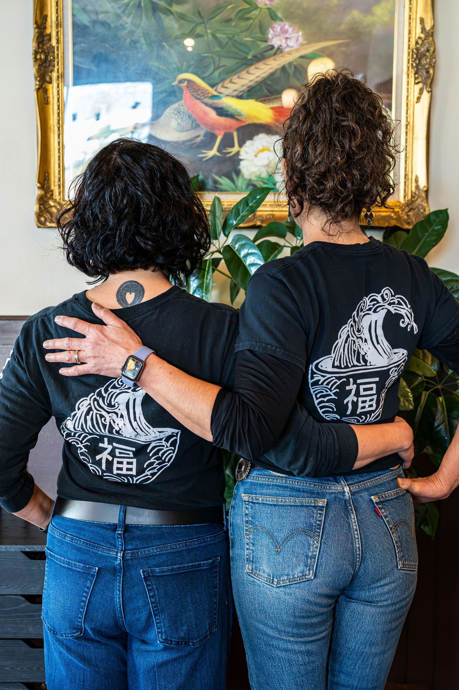 Two women wearing matching black long-sleeve shirts with Japanese wave bowl graphic stand with arms around each other.