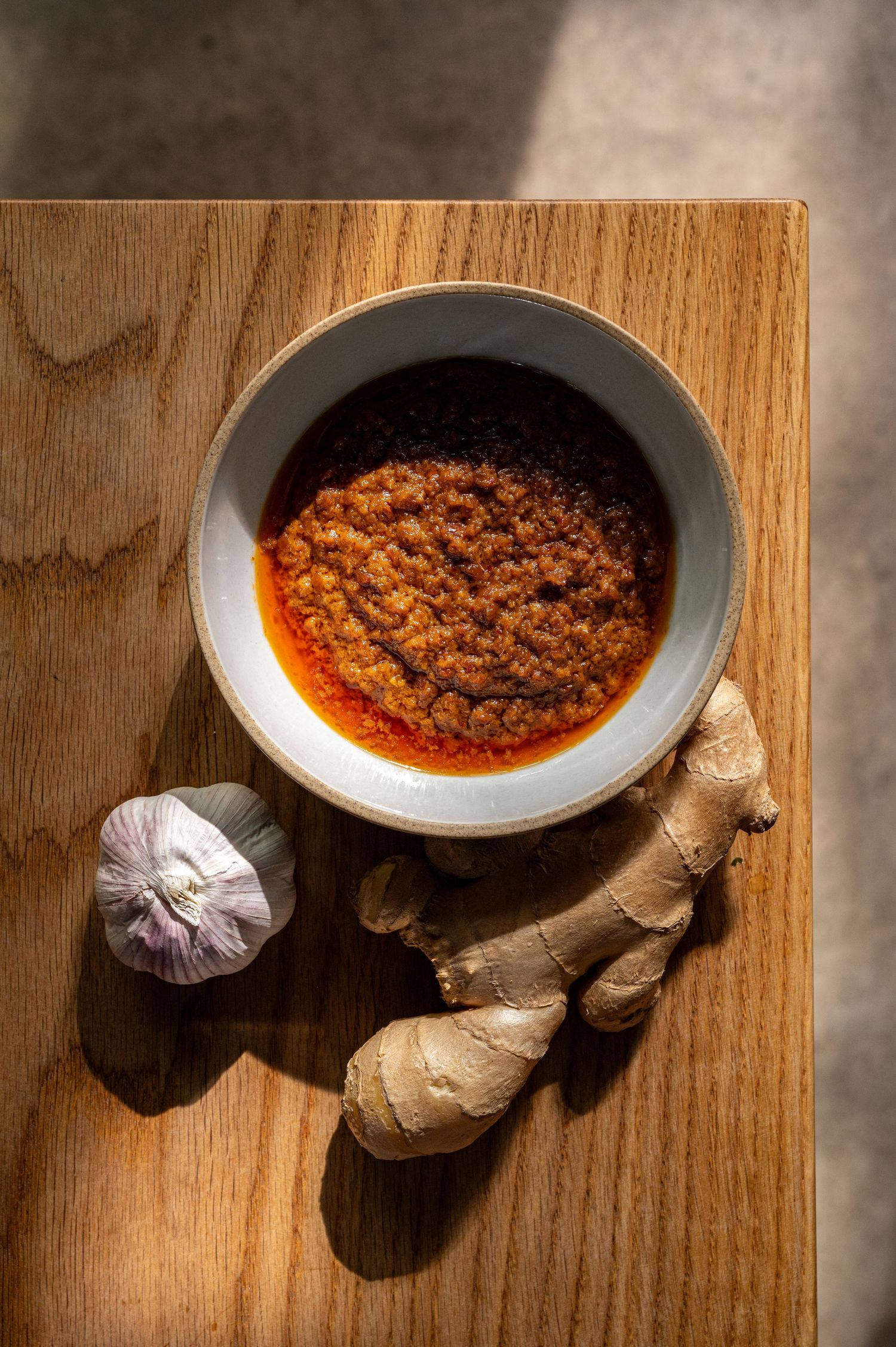 A bowl of red chili paste on a wooden board with fresh ginger root and garlic bulb beside it.