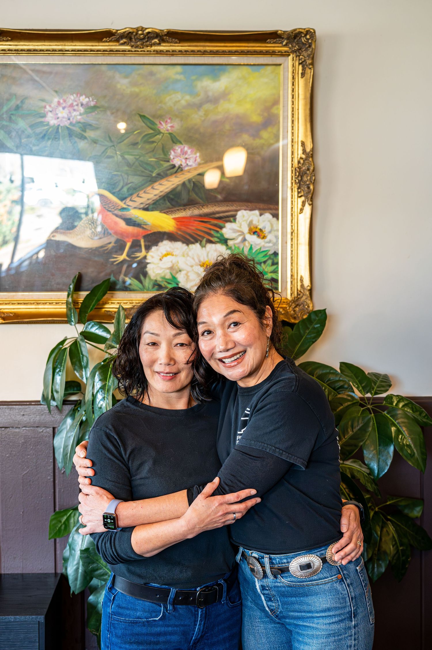 Two women in black shirts embrace warmly in front of a ornate gold-framed painting with tropical birds.