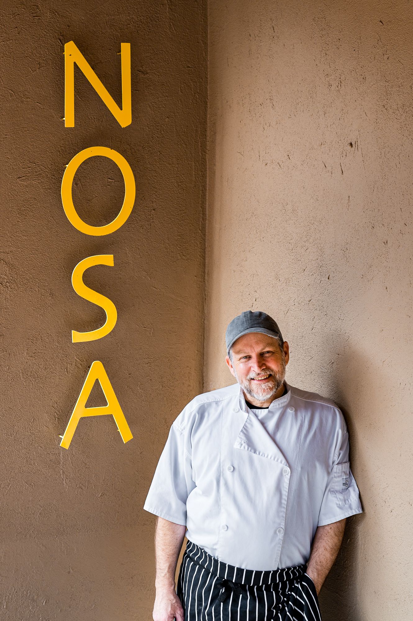 Chef in white coat smiling beside yellow NOSA restaurant sign on textured wall.