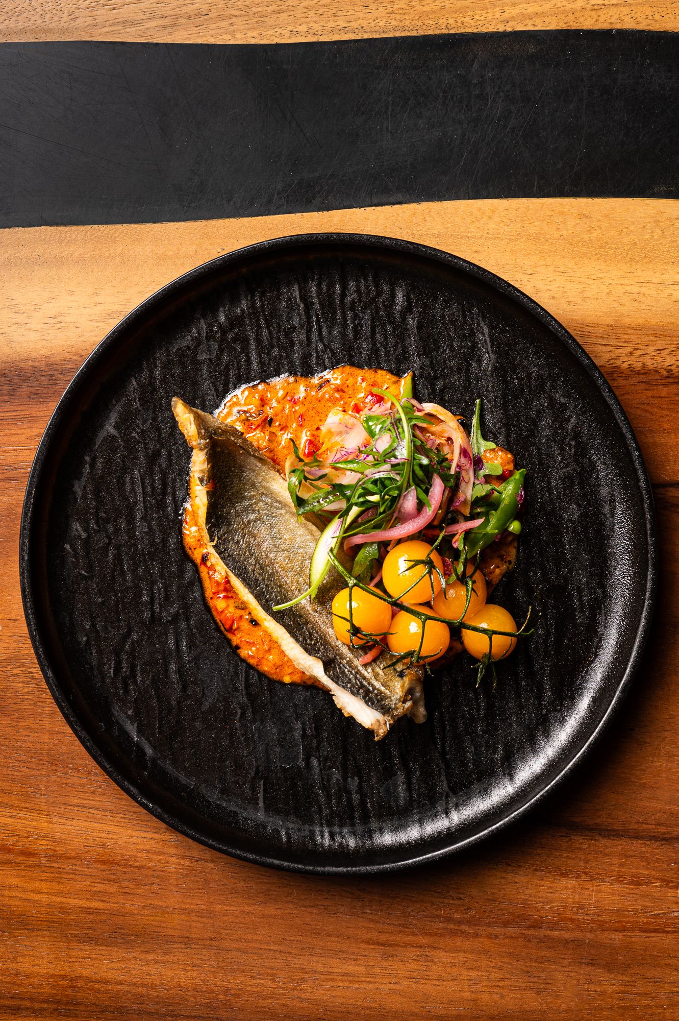 Elegant plated fish dish with roasted tomato sauce, golden cherry tomatoes, pickled red onions, and microgreens on a black ceramic plate.