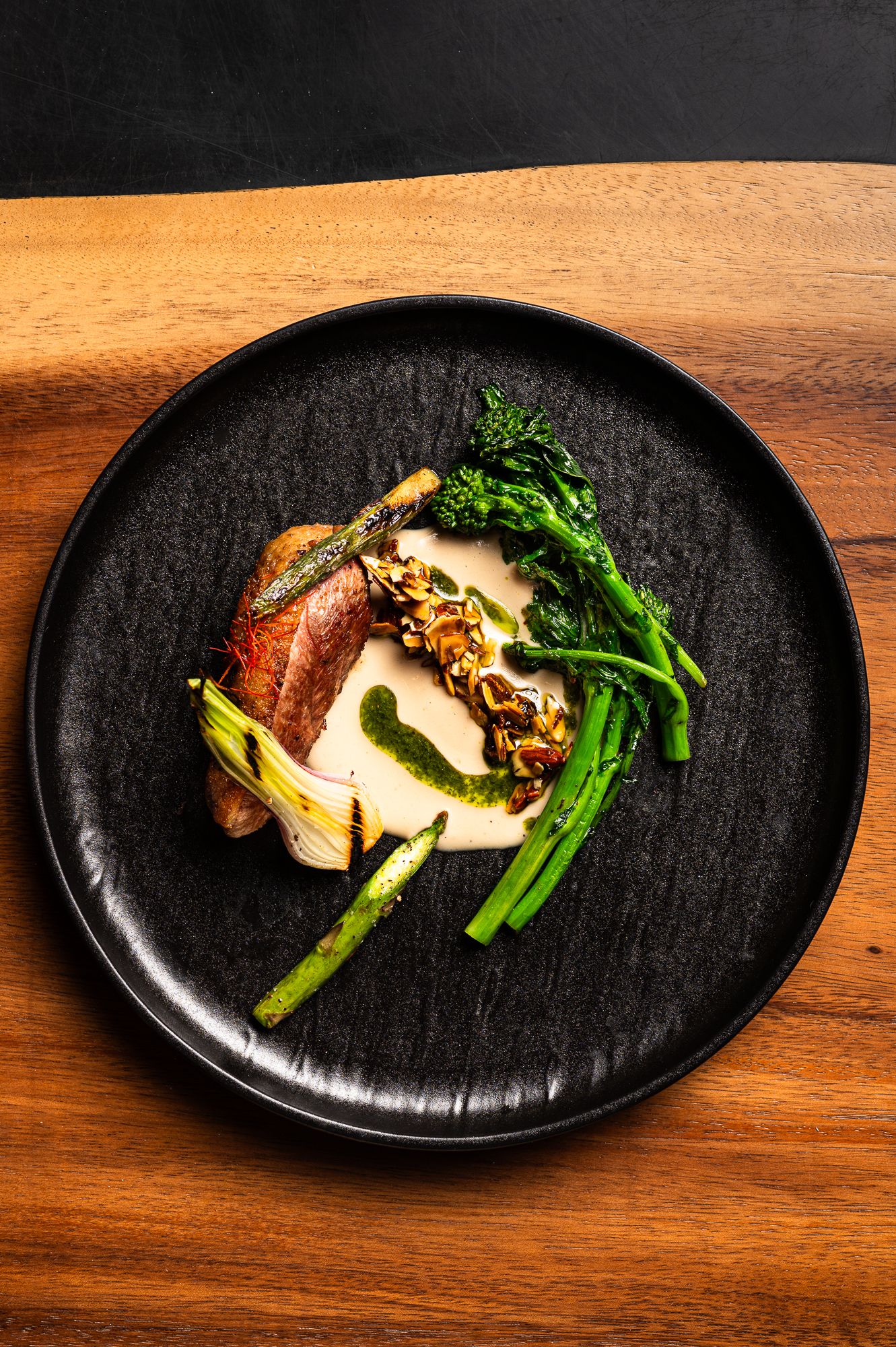 Elegant fine dining plate with seared meat, broccolini, asparagus, mushrooms, and cream sauce on a black ceramic plate.