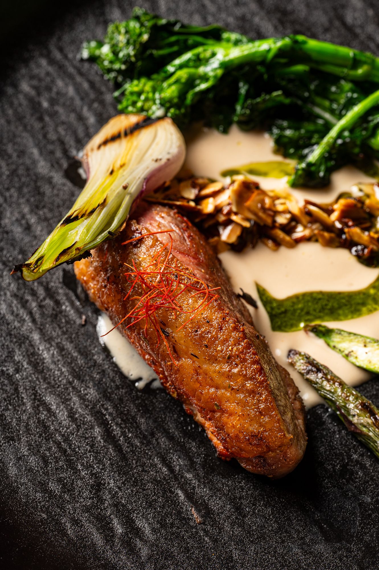 Elegant plated duck breast with broccolini, mushrooms, and cream sauce on slate, garnished with saffron threads.
