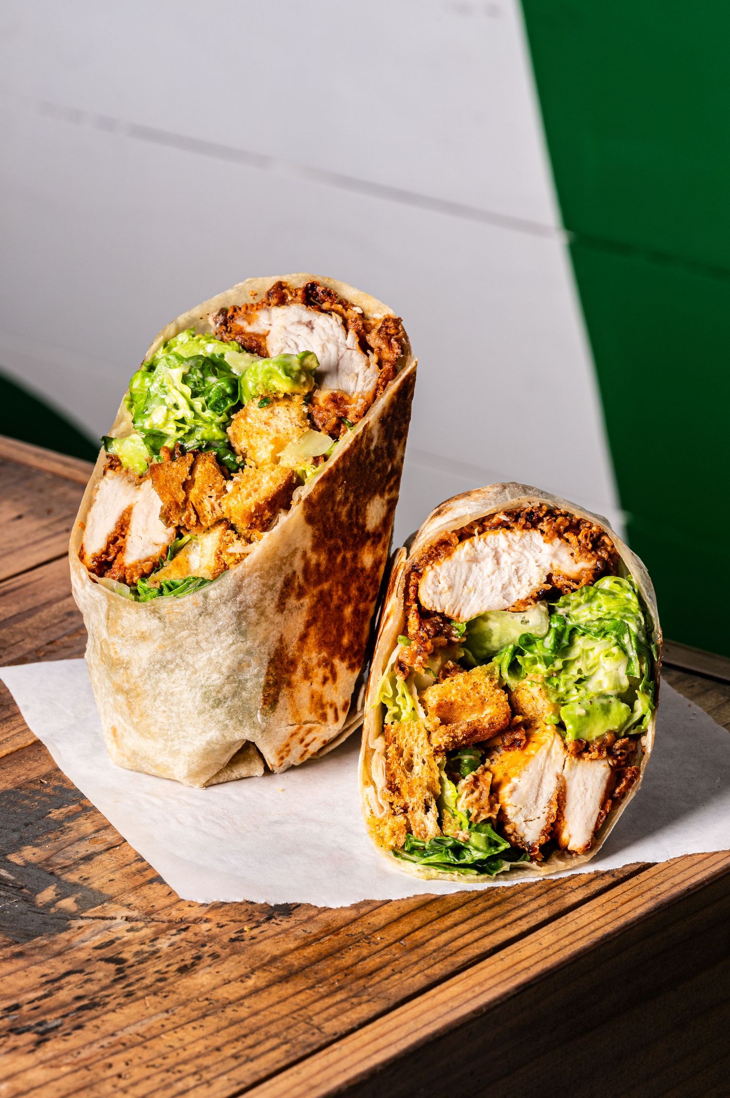 A halved chicken burrito with crispy coating, lettuce, and cheese on parchment paper atop a wooden surface.