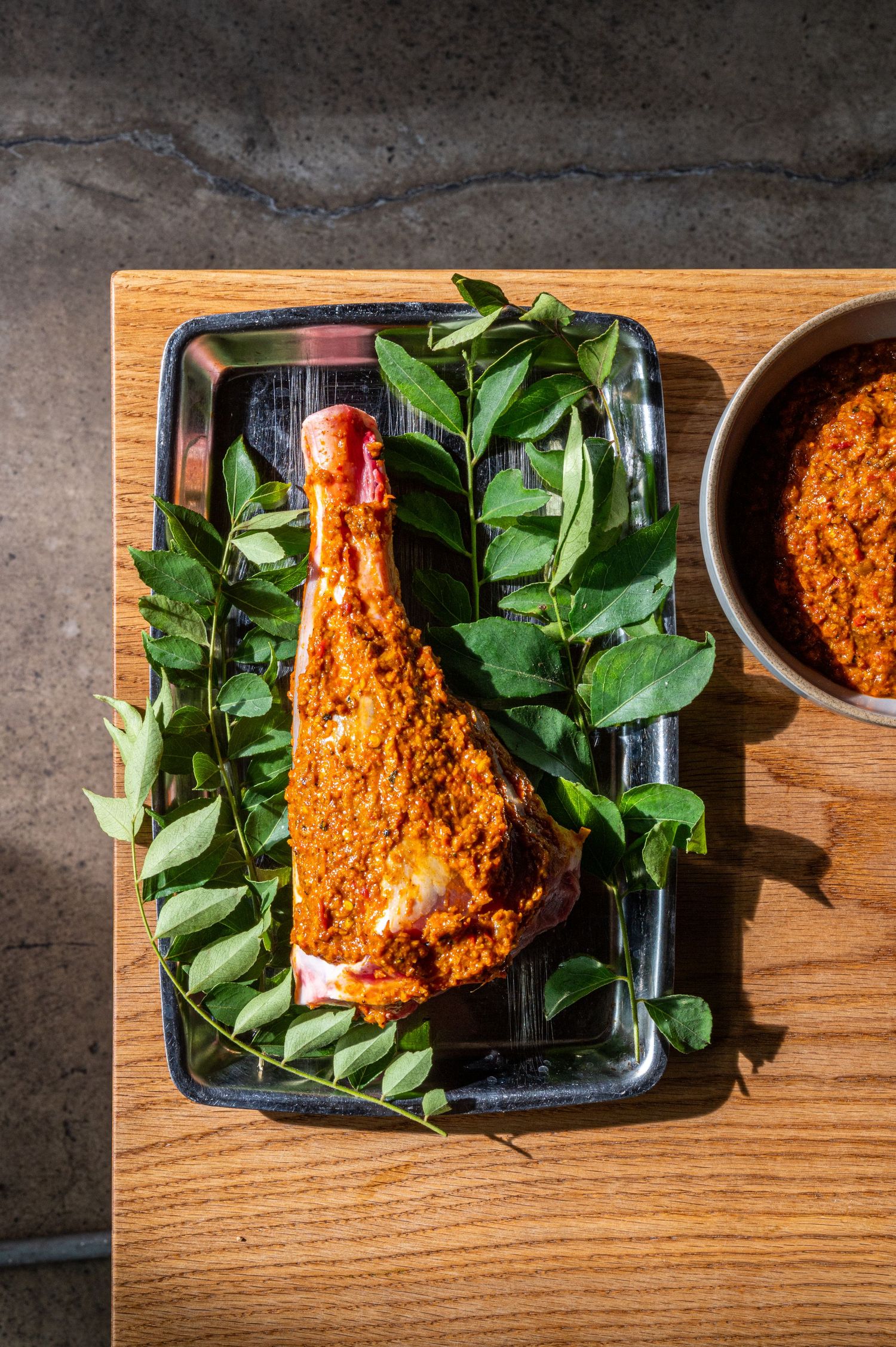 Spiced chicken leg on fresh curry leaves in a dark tray beside a bowl of red spice paste on a wooden board.