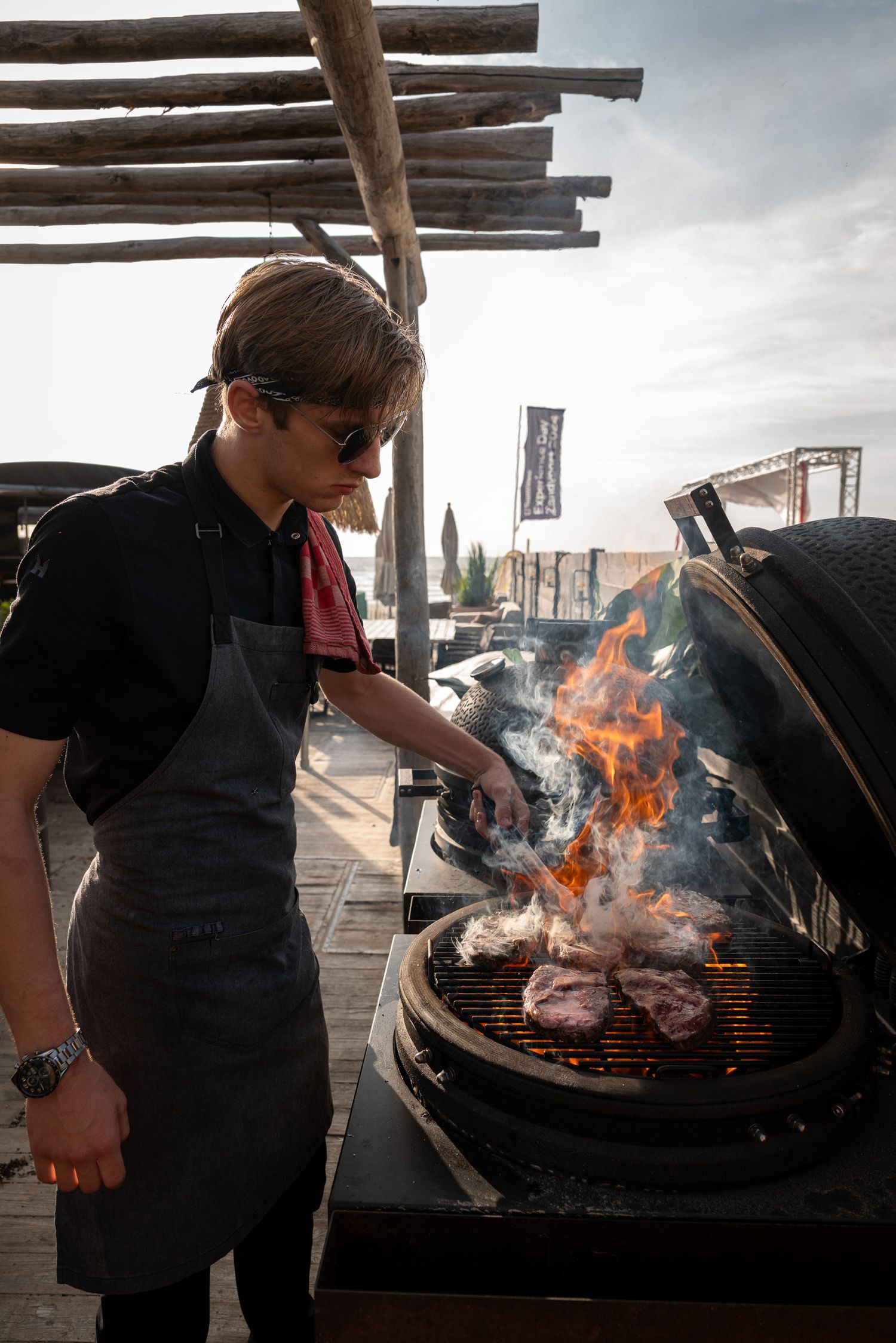 Chef grilling food over open flames on an outdoor barbecue with smoke rising.