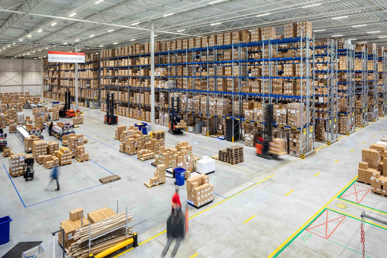 Large warehouse interior with tall storage racks, cardboard boxes, and workers operating equipment on the floor.