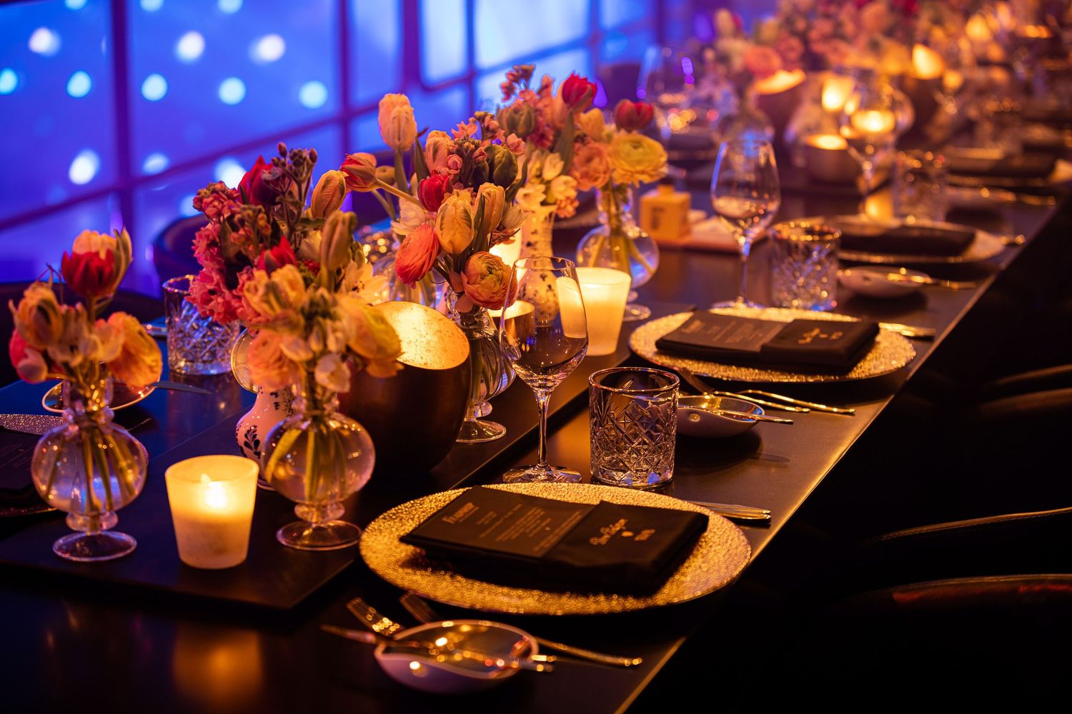Elegant dinner table setting with glowing candles and orange floral centerpieces in glass vases against blue lighting.