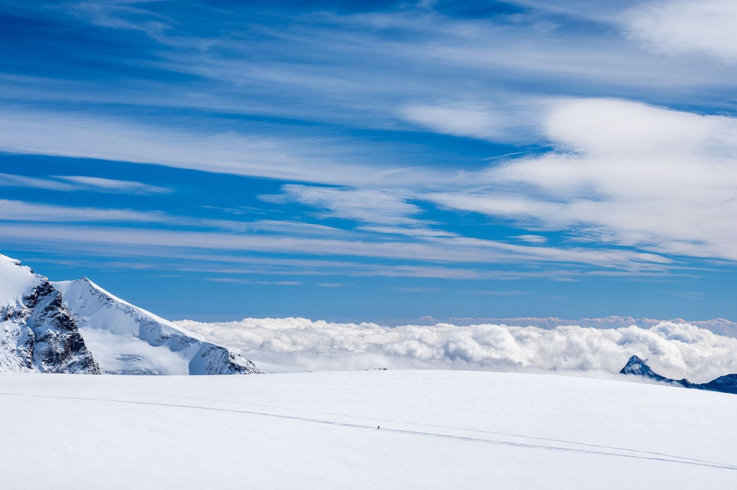 Expansive view of snow-covered mountain peaks against bright blue sky with wispy clouds.