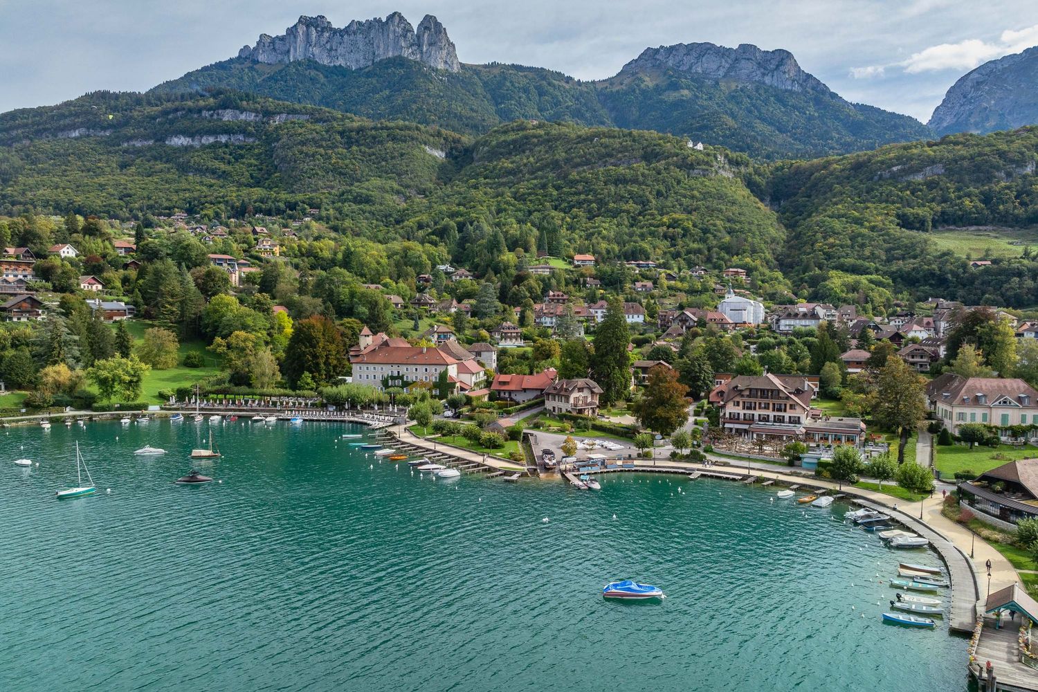 Aerial view of a lakeside village nestled between mountains with boats dotting the turquoise waters.