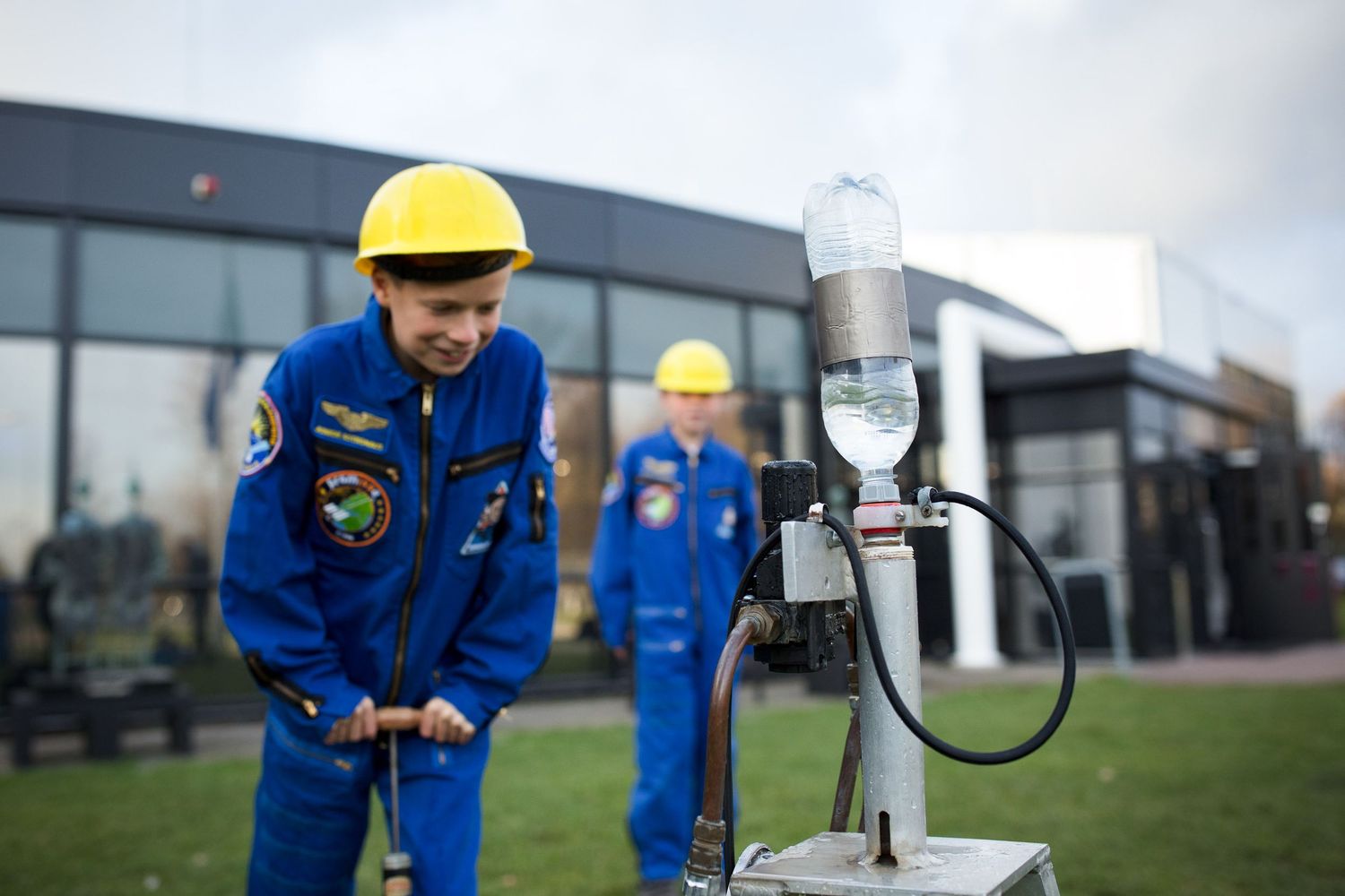 Children in blue jumpsuits and yellow hard hats conduct a science experiment with a water rocket outdoors.