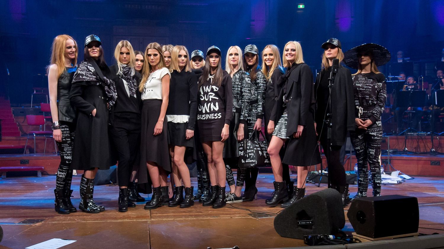 A group of female models posing together on a stage during a fashion show wearing edgy dark outfits.
