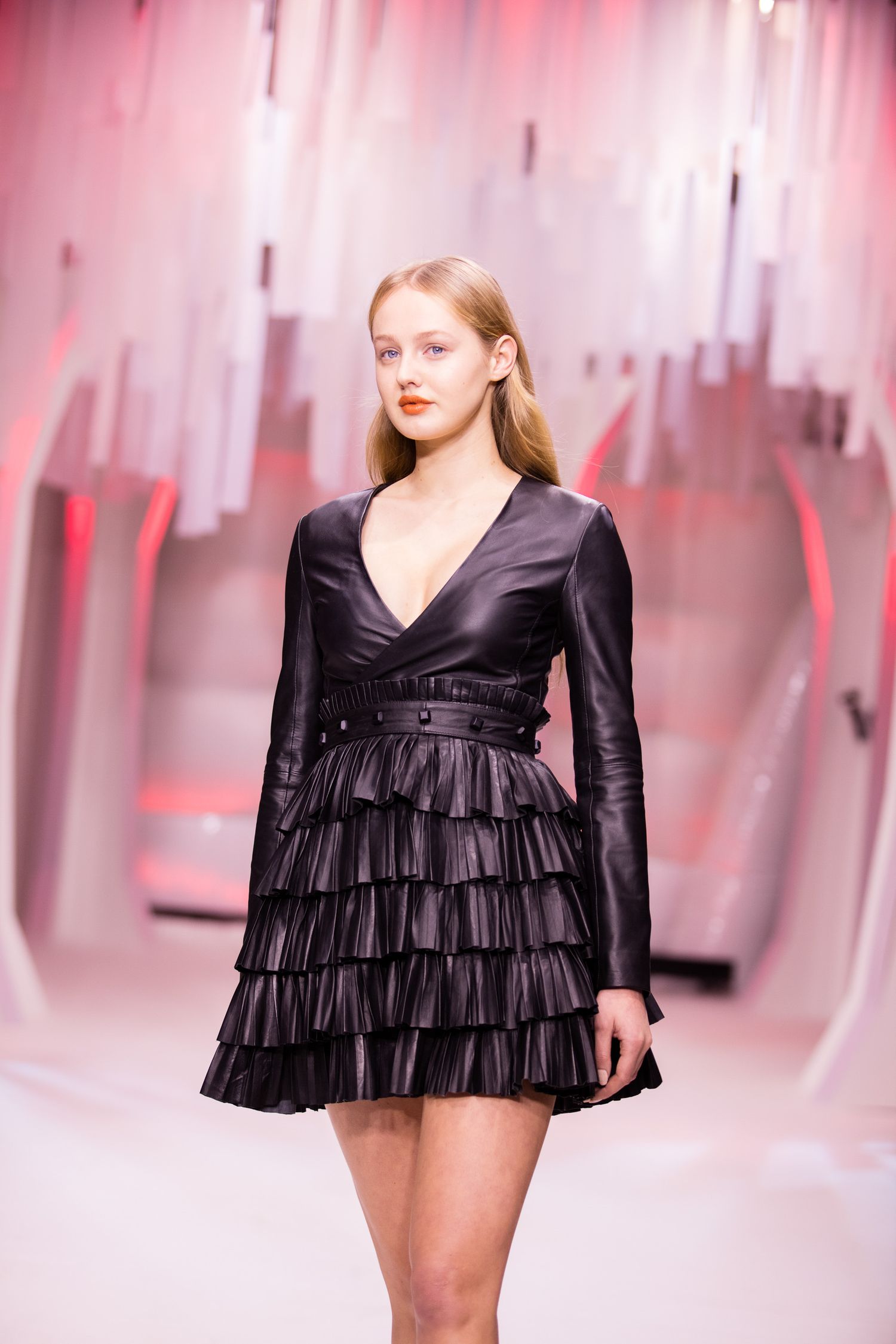Model walks runway in black leather tiered ruffle mini dress with V-neckline and long sleeves at fashion show.