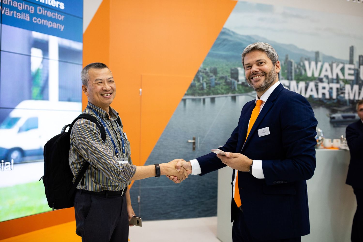 Two professionals shake hands in front of a corporate display with orange and blue design elements.