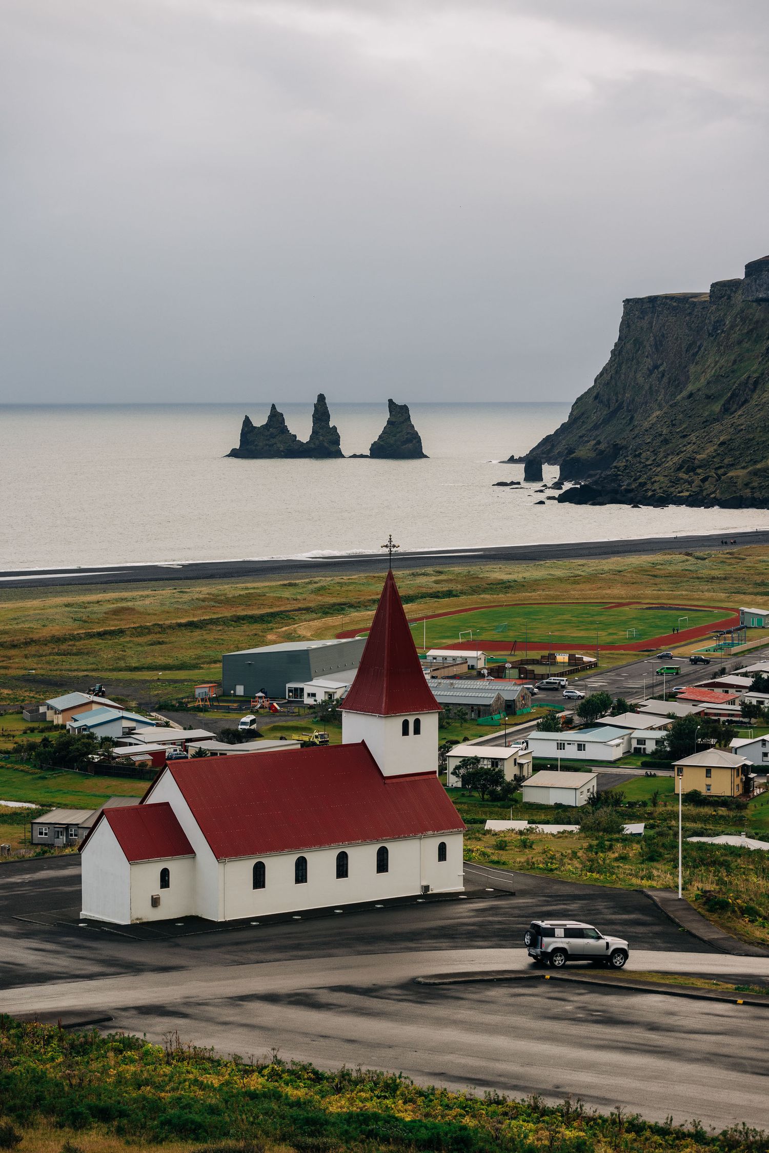 White church with red roof overlooks coastal village and dramatic rock formations in Vik, Iceland against moody gray sky.
