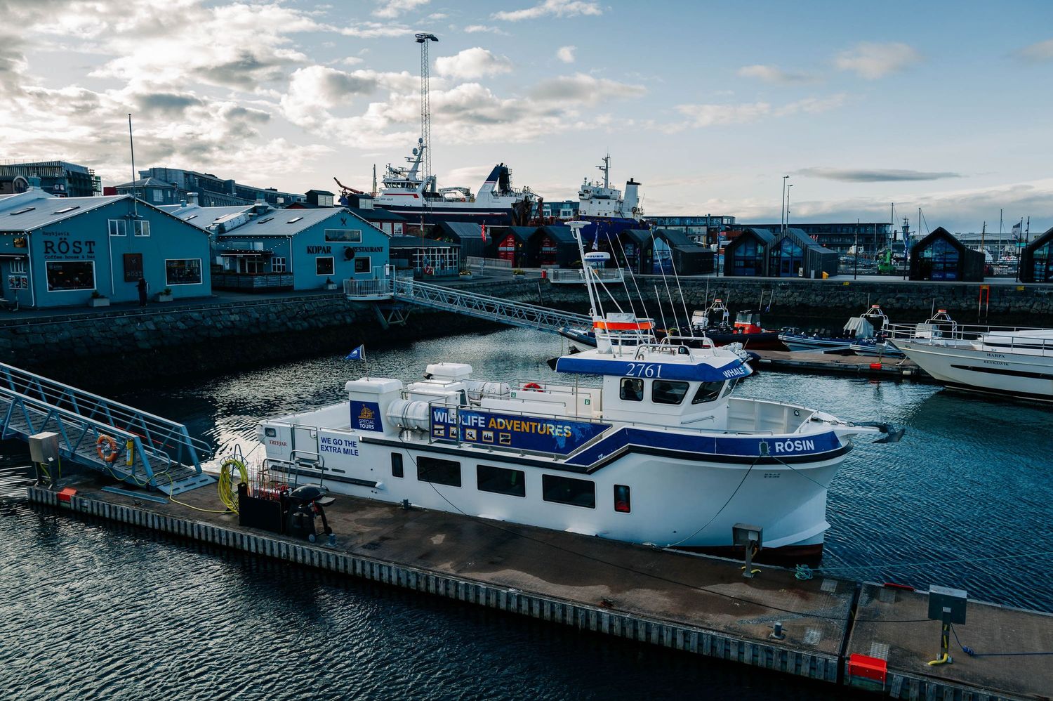 Fishing boats docked at a harbor during sunset with blue buildings and industrial structures along the waterfront.