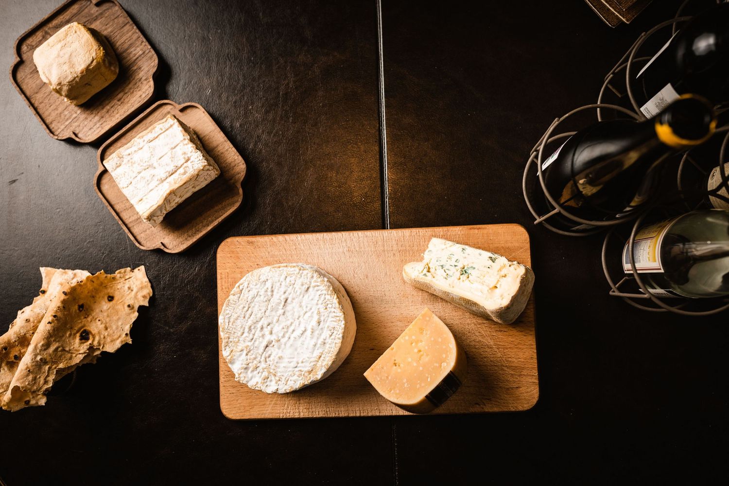 Various artisanal cheeses displayed on rustic wooden cutting boards with wine bottles in dark ambient setting.