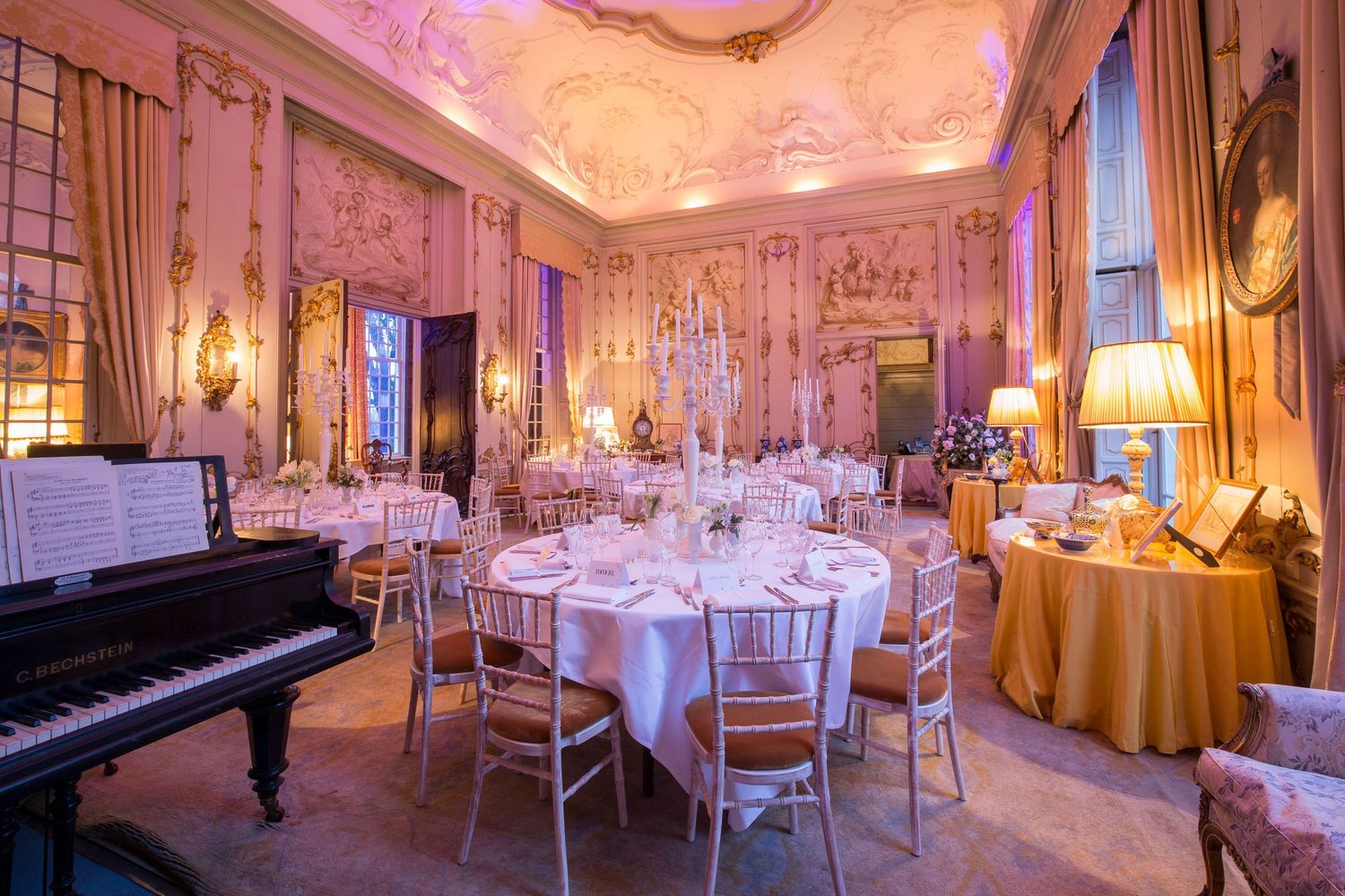 Elegant banquet hall with round tables, chiavari chairs, grand piano, ornate pink walls, and warm romantic lighting.